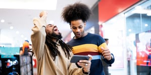 one man celebrates something on his mobile phone while another looks on